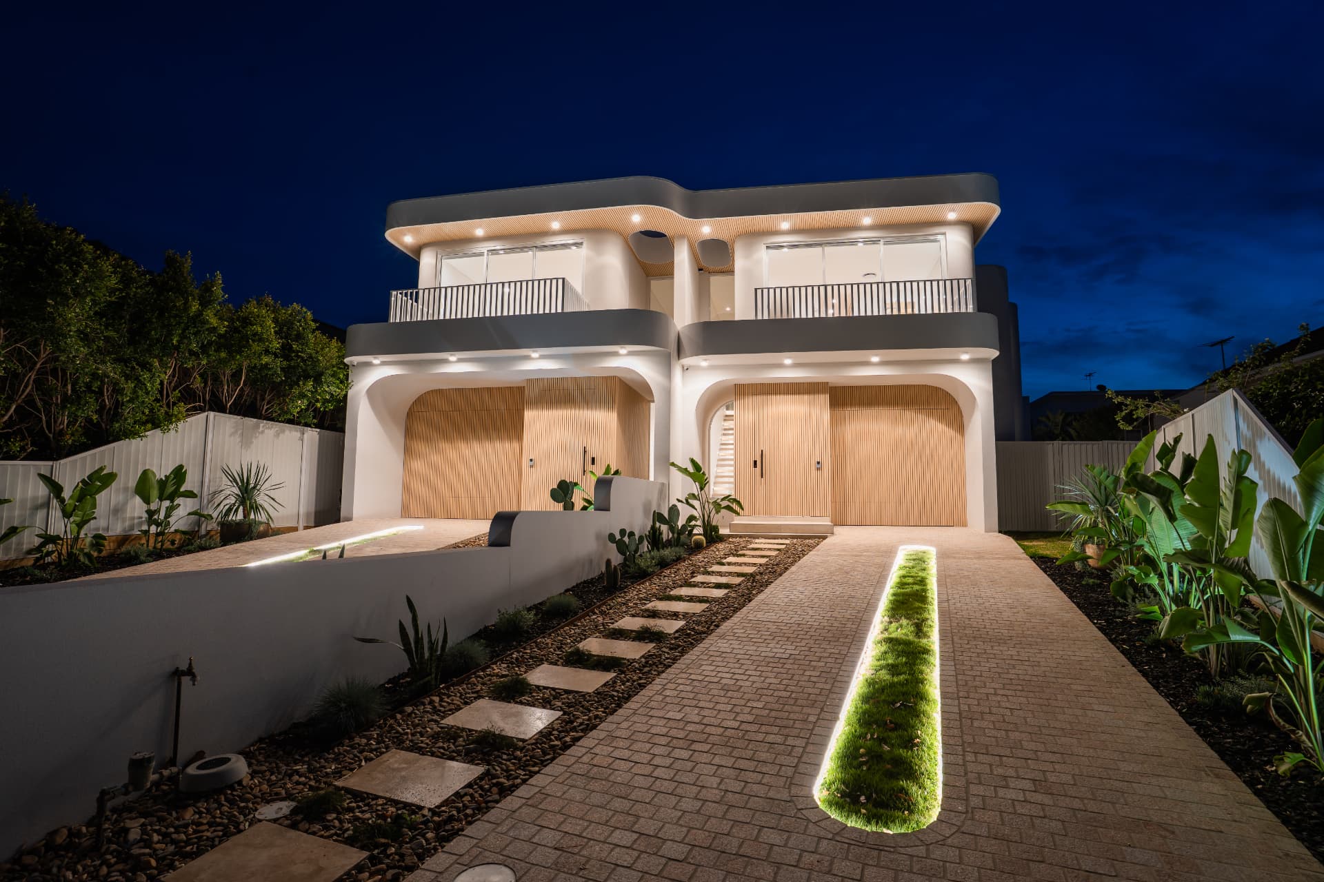The Cronulla duplex at dusk with facade downlights, lit driveway strip and timber garage doors glowing