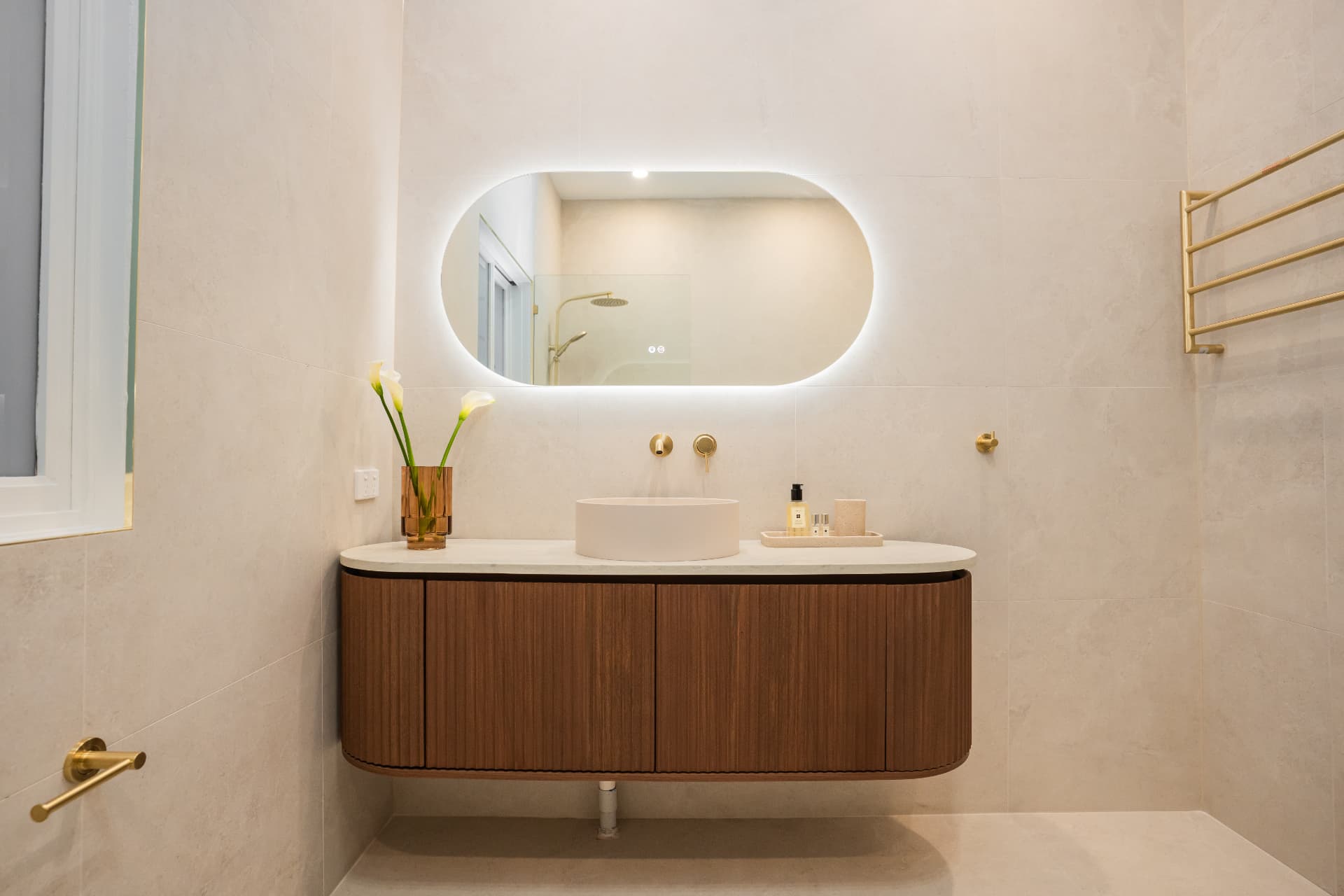 Fluted timber vanity with stone top, round basin, brushed brass tapware and a backlit oval mirror