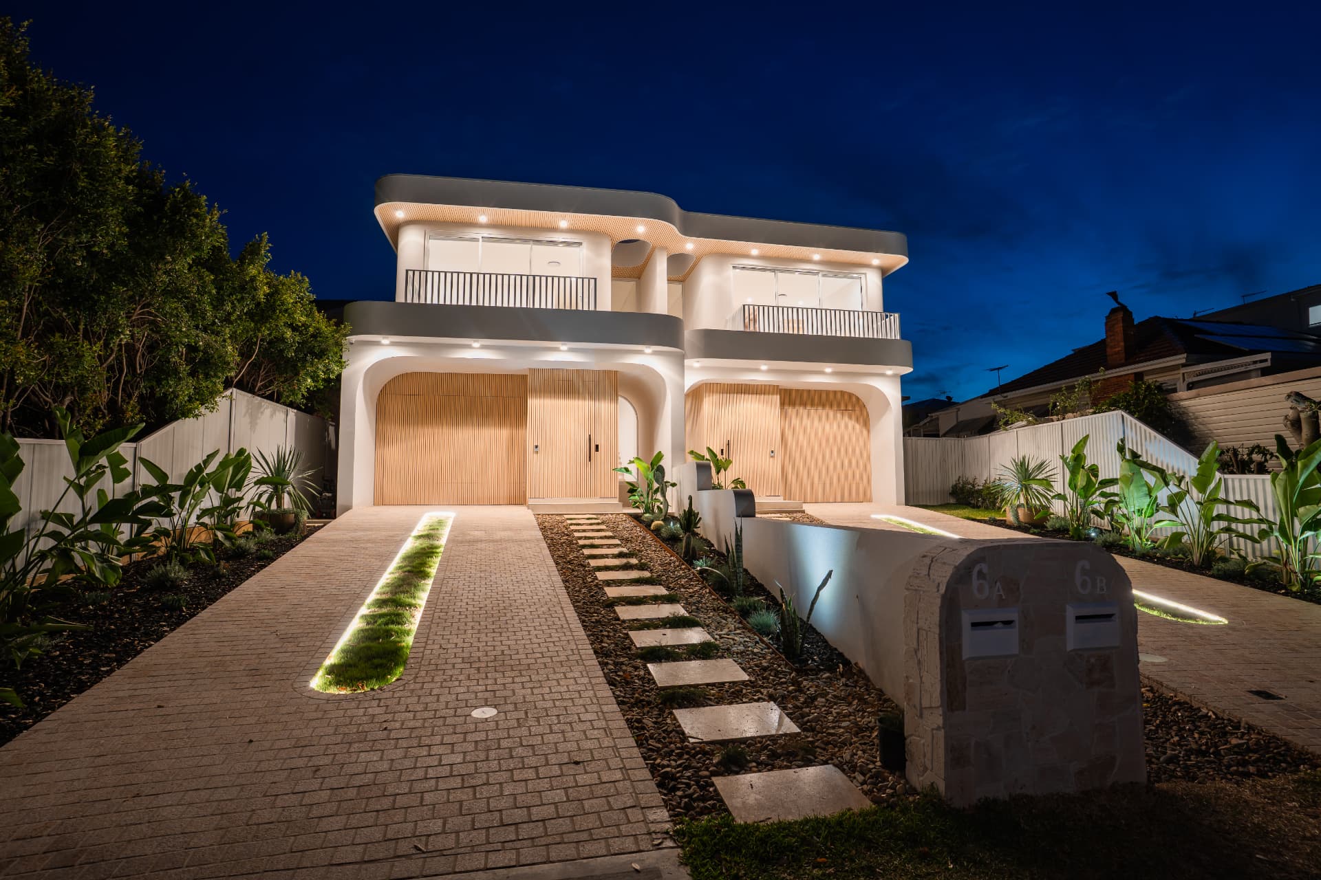Dusk view of the lit duplex facade with timber garage doors, stepping-stone path and sandstone letterbox