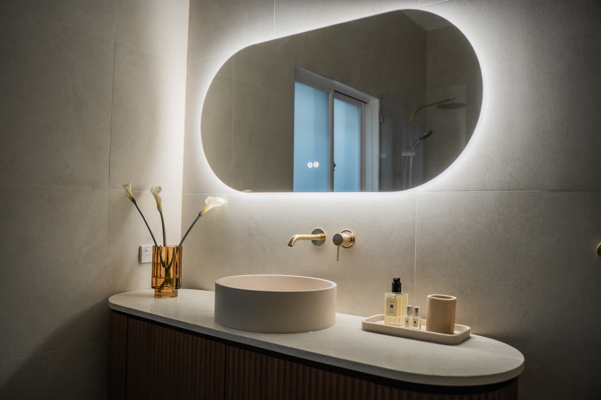 Detail of the vanity with brushed brass wall mixer and backlit mirror glow on large-format tiles